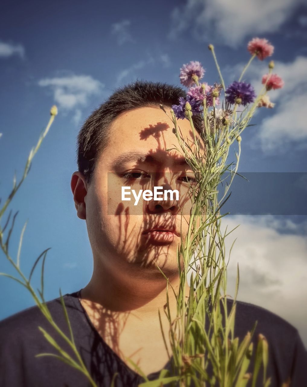 Close-up of young man with cornflowers against cloudy blue sky.