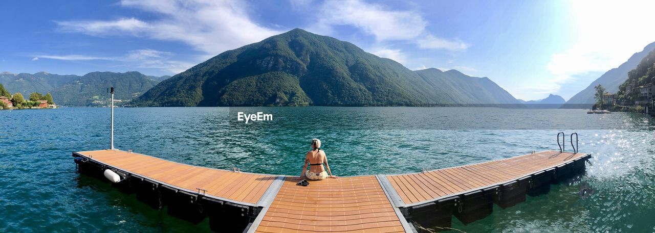 Rear view of woman sitting on pier over sea against mountains and sky