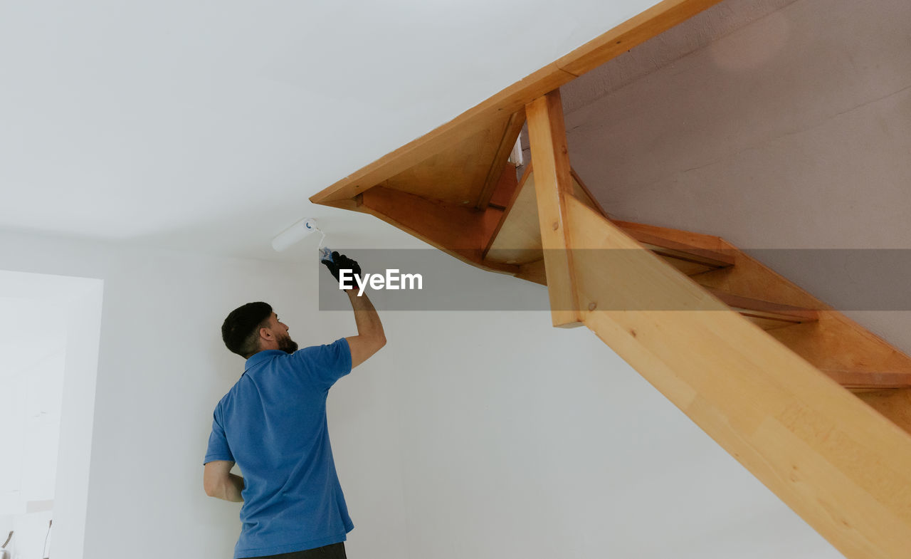 A young oriental man paints the ceiling under the stairs with a roller.