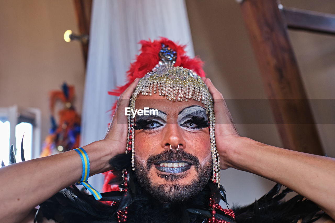 Cheerful adult bearded drag queen male with bright makeup wearing extravagant costume with feathers adjusting headdress while preparing for lgbt performance
