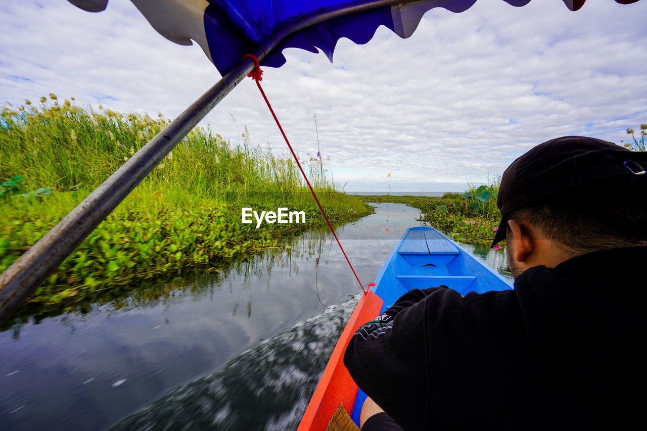 REAR VIEW OF MAN SITTING ON RIVERBANK AGAINST SKY