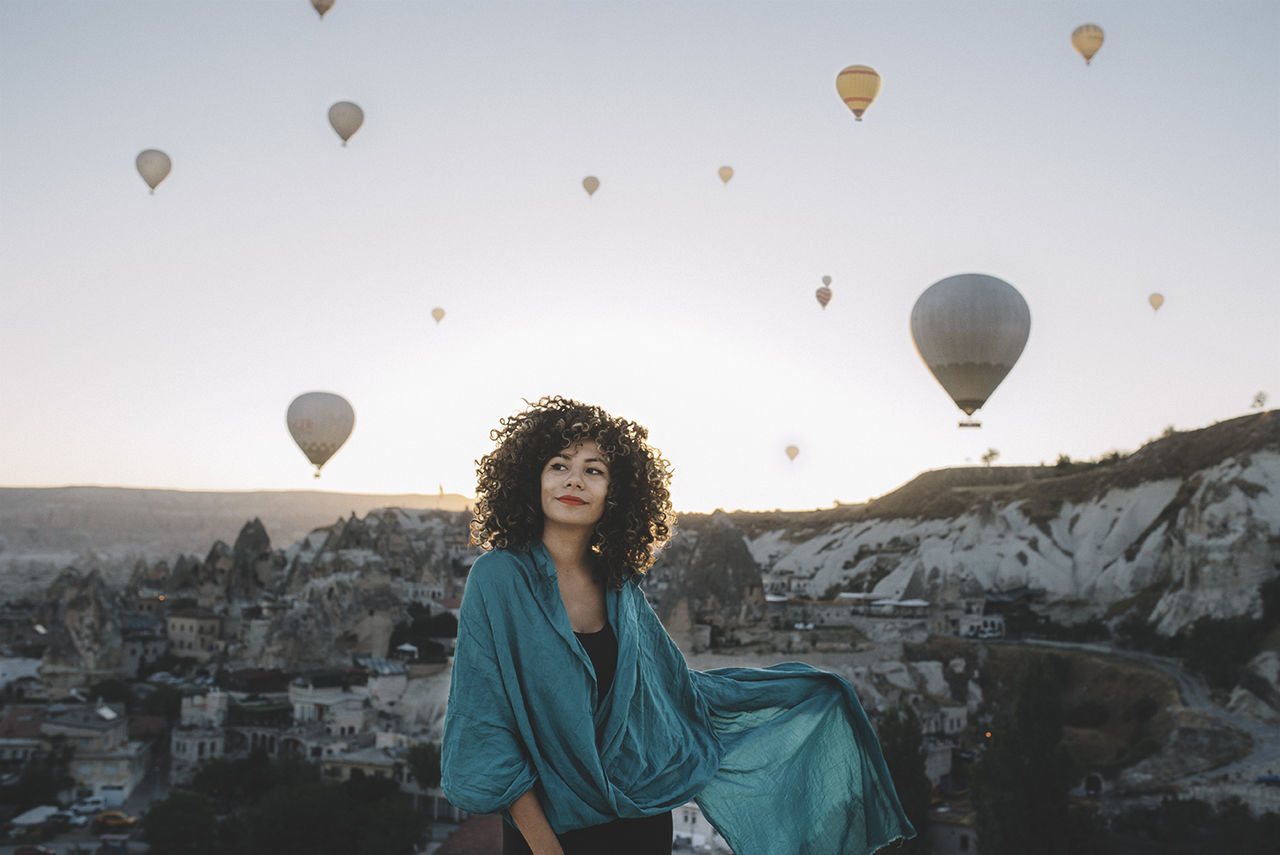 Young woman looking away while standing against hot air balloons flying in sky