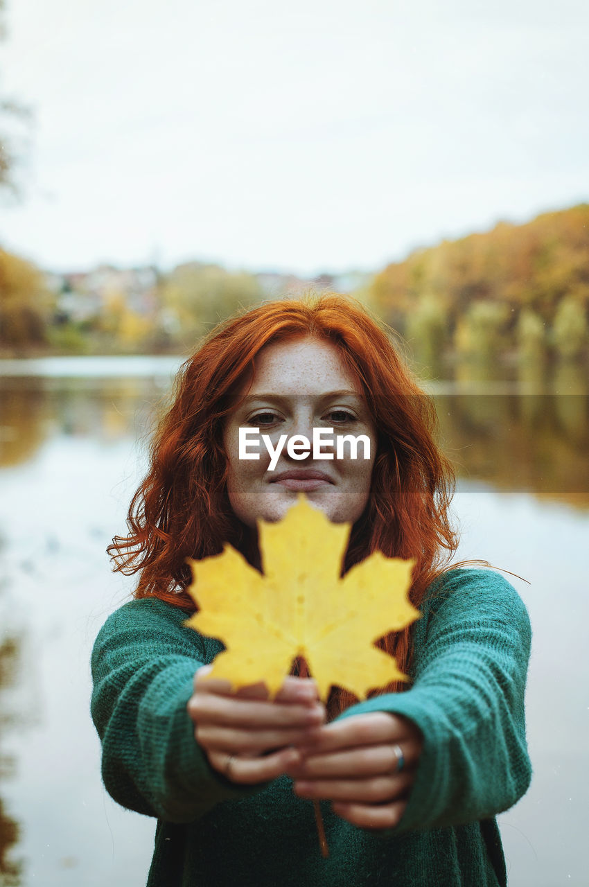 Redhead funny woman walking with yellow leaf near lake in autumn park.