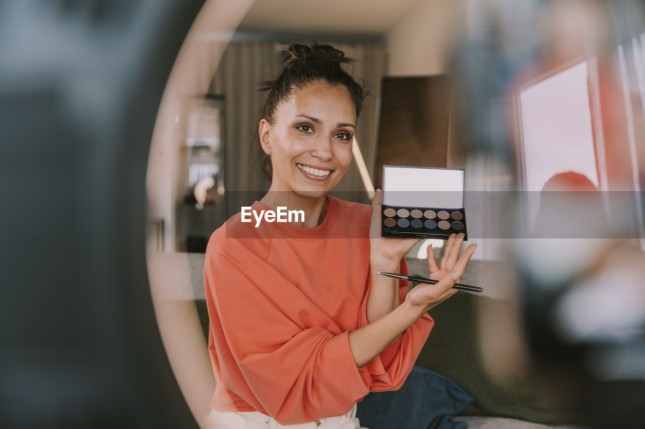 Smiling female influencer showing make-up palette while doing tutorial at home