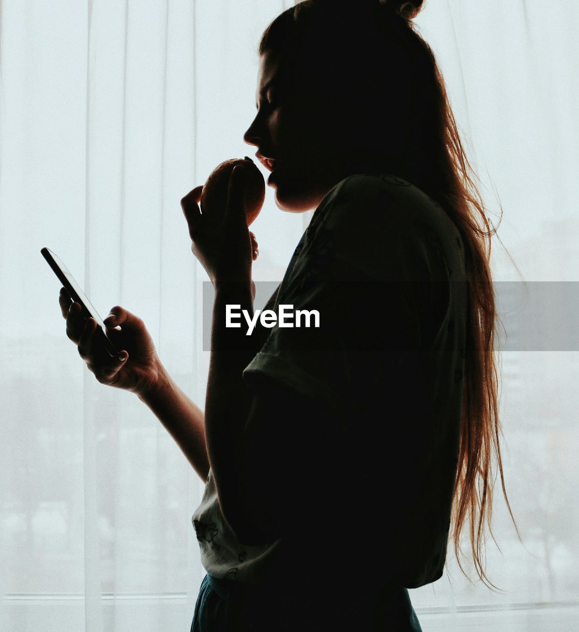 Side view of young woman using smart phone while standing against window