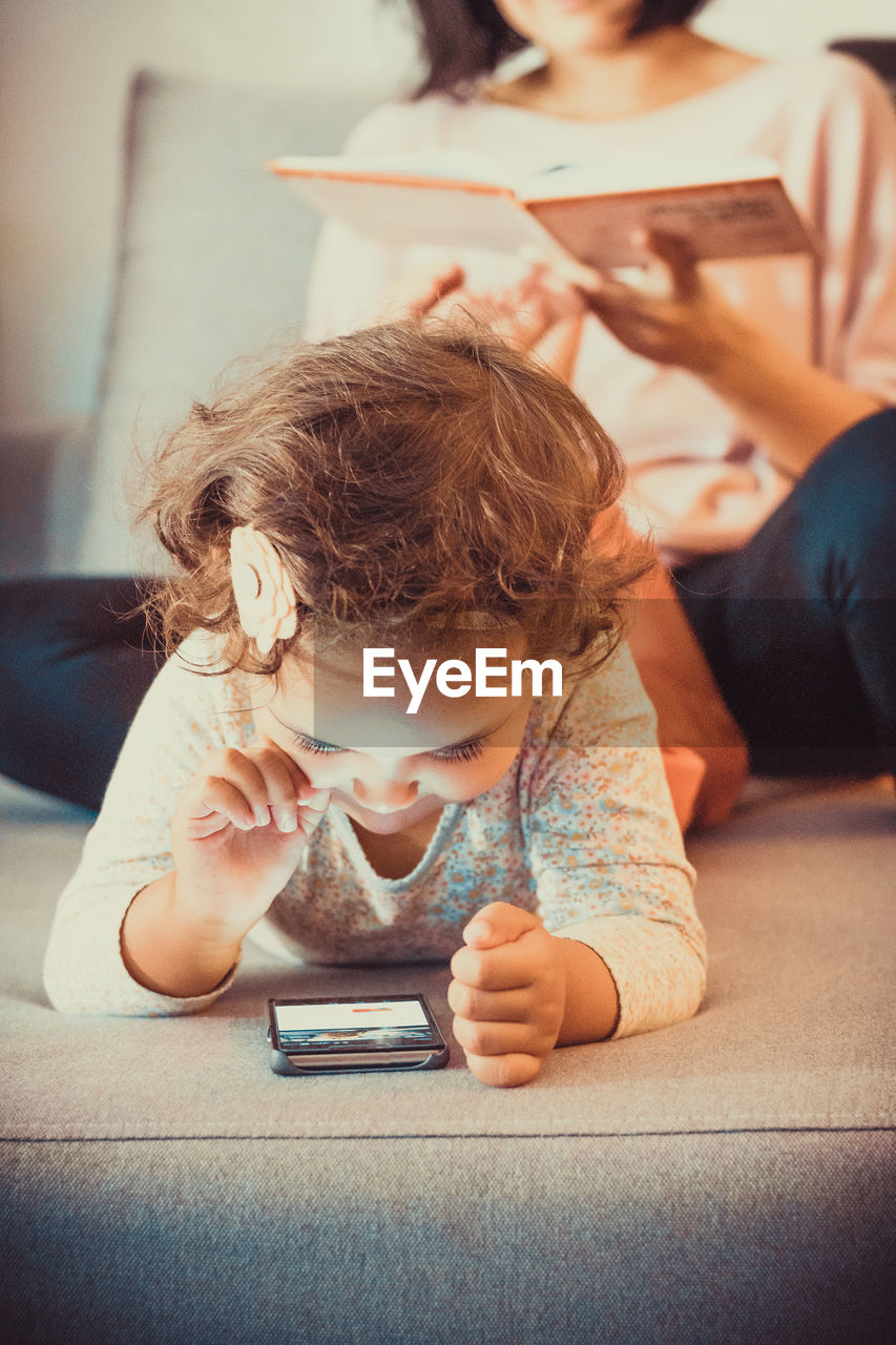 Cute small girl surfing the net on smart phone while relaxing at home with her mother.