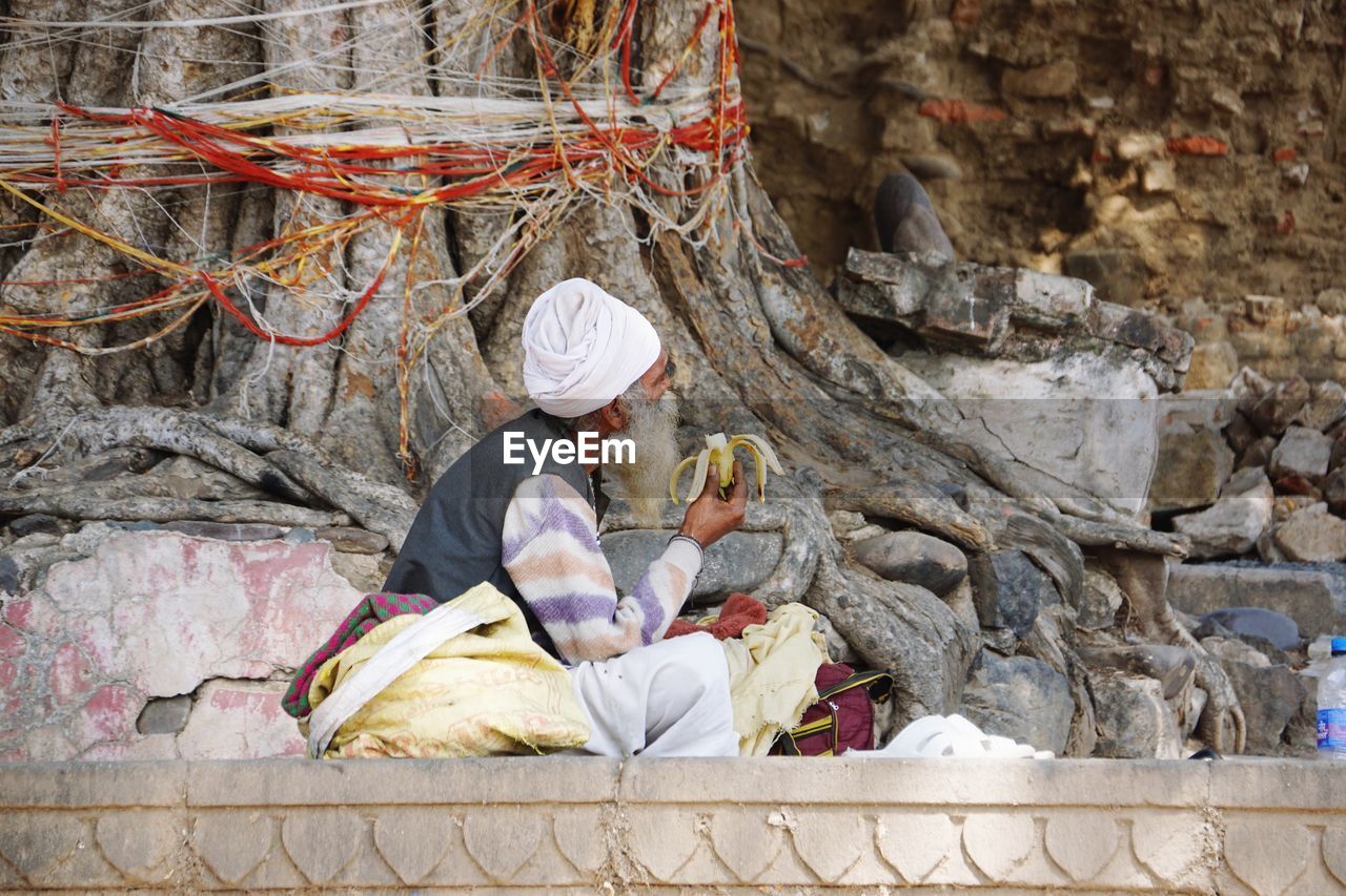 Side view of man eating banana while sitting against tree trunk