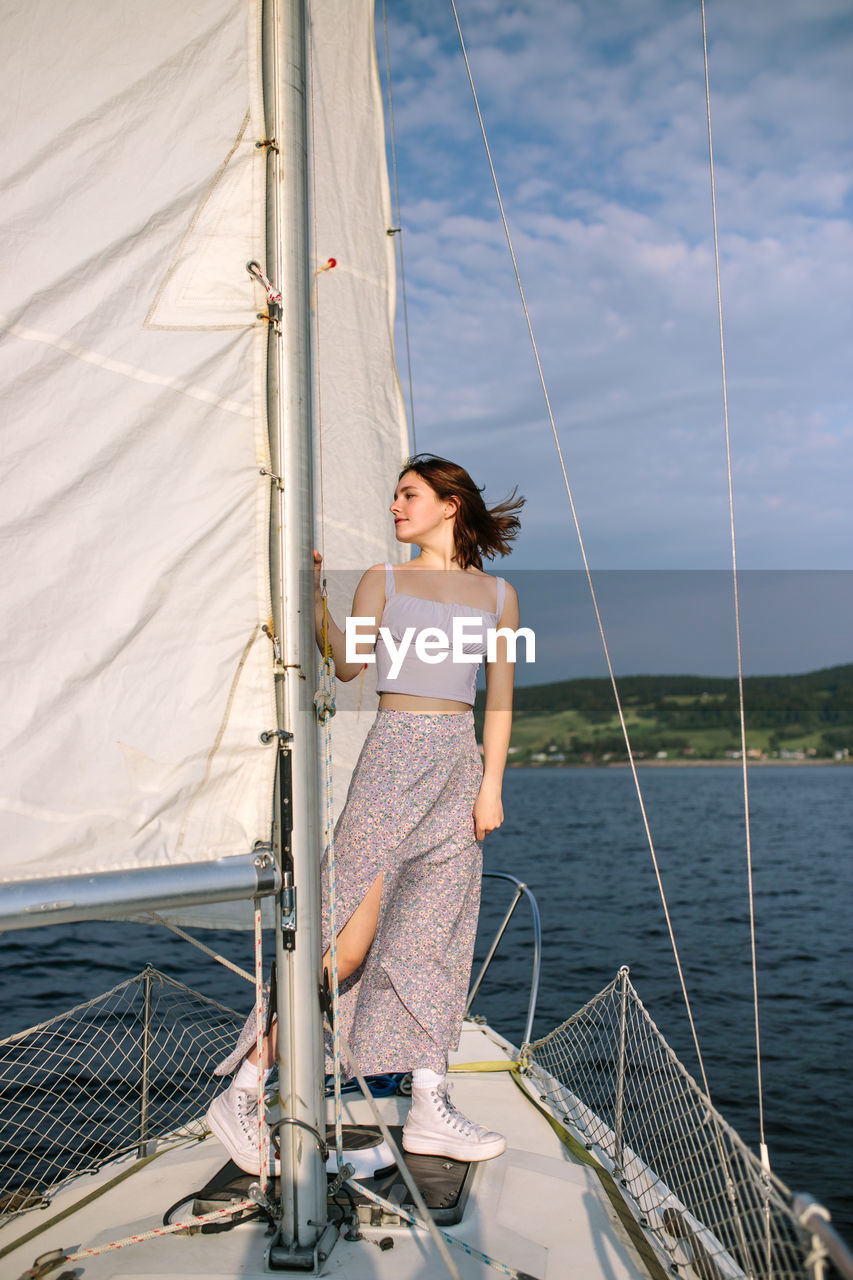 Full body of trendy young female tourist in stylish outfit standing on deck of modern yacht sailing in sea and looking away during summer holidays