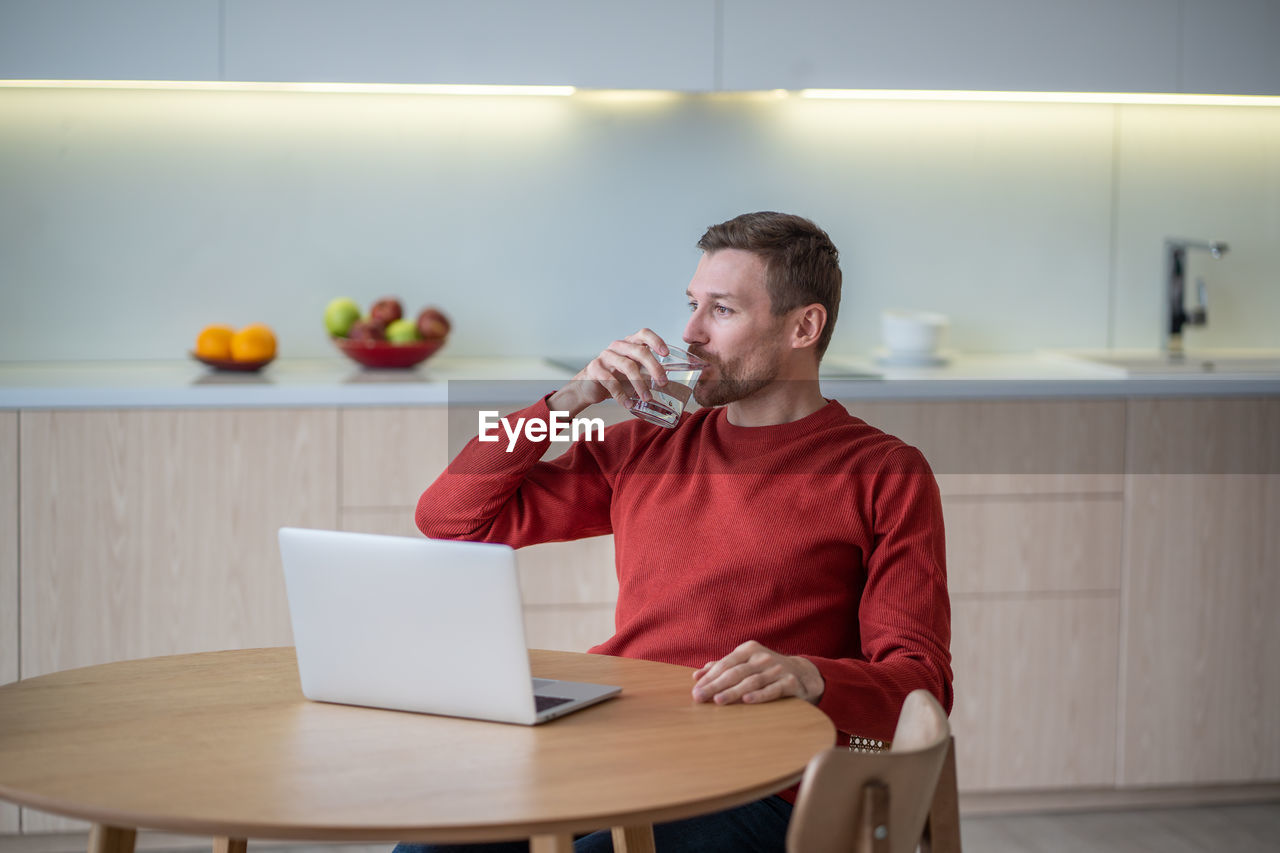 Freelancer man drinking water having break in work sitting at table on kitchen at home with laptop.