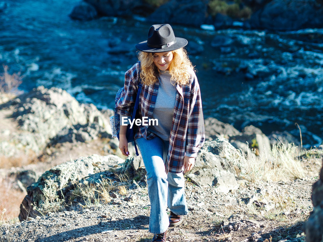 Young curly woman in hat and plaid t-shirt with backpack hiking on mountain with river view