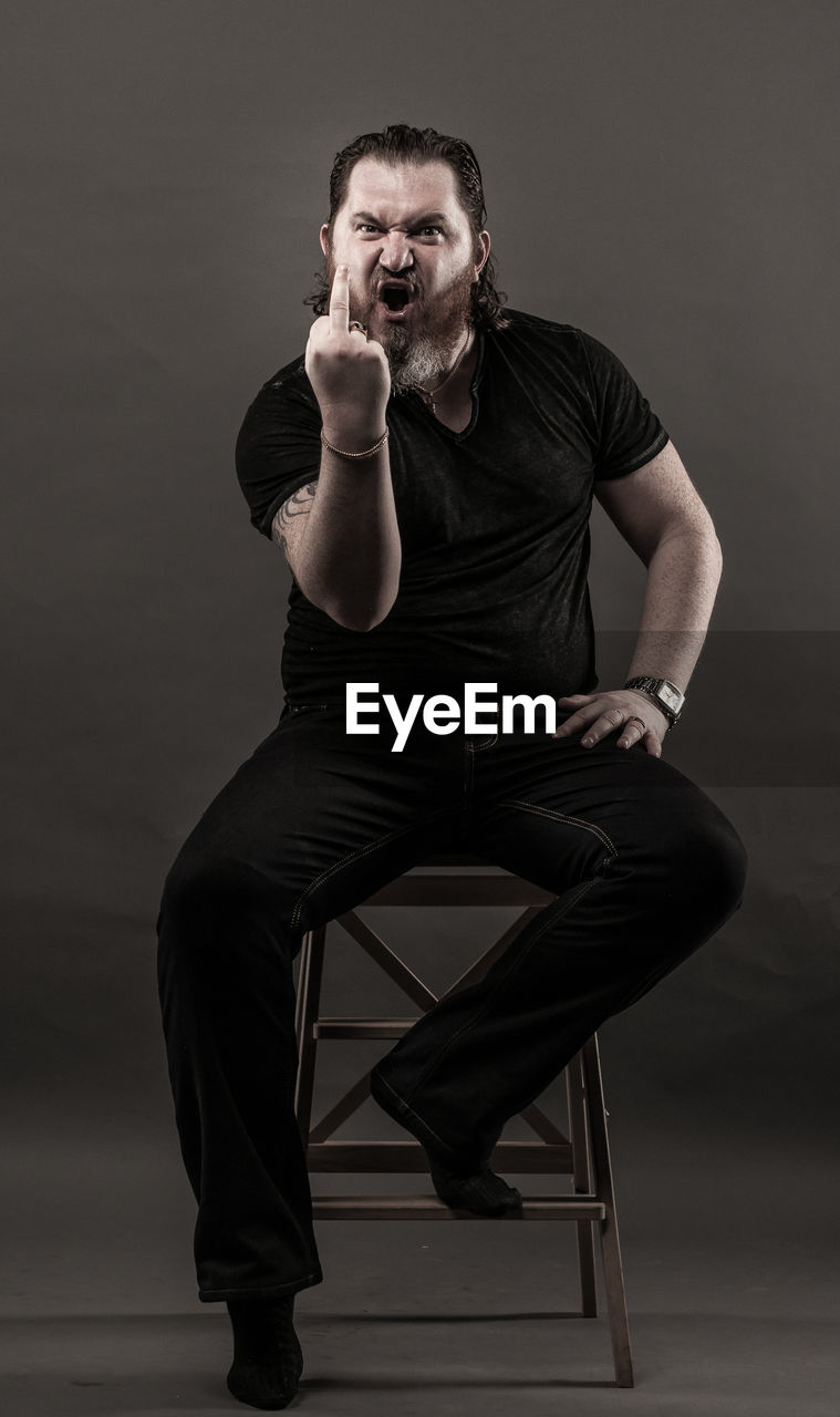 Mid adult man gesturing obscene gesture while sitting against black background