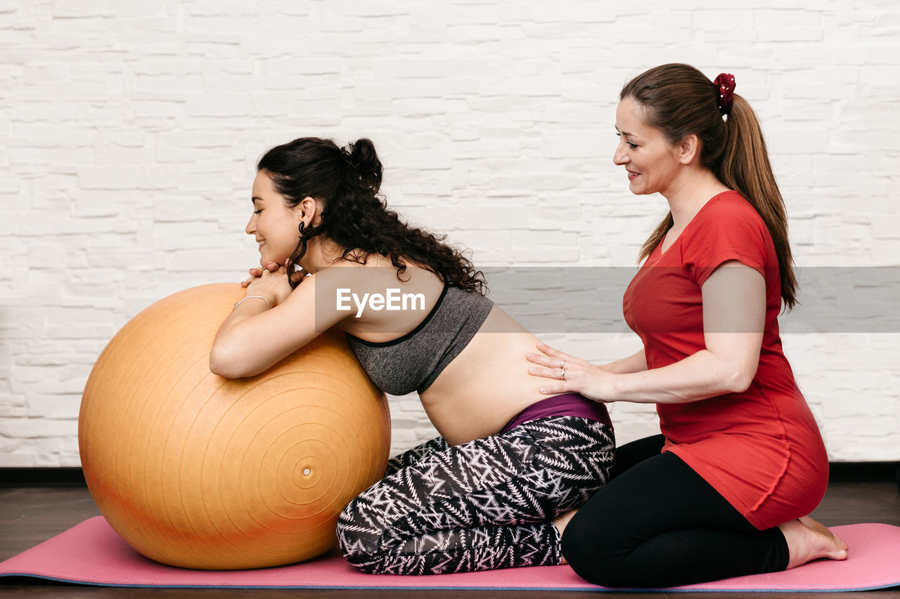 Fitness instructor assisting pregnant woman while exercising with fitness ball at home
