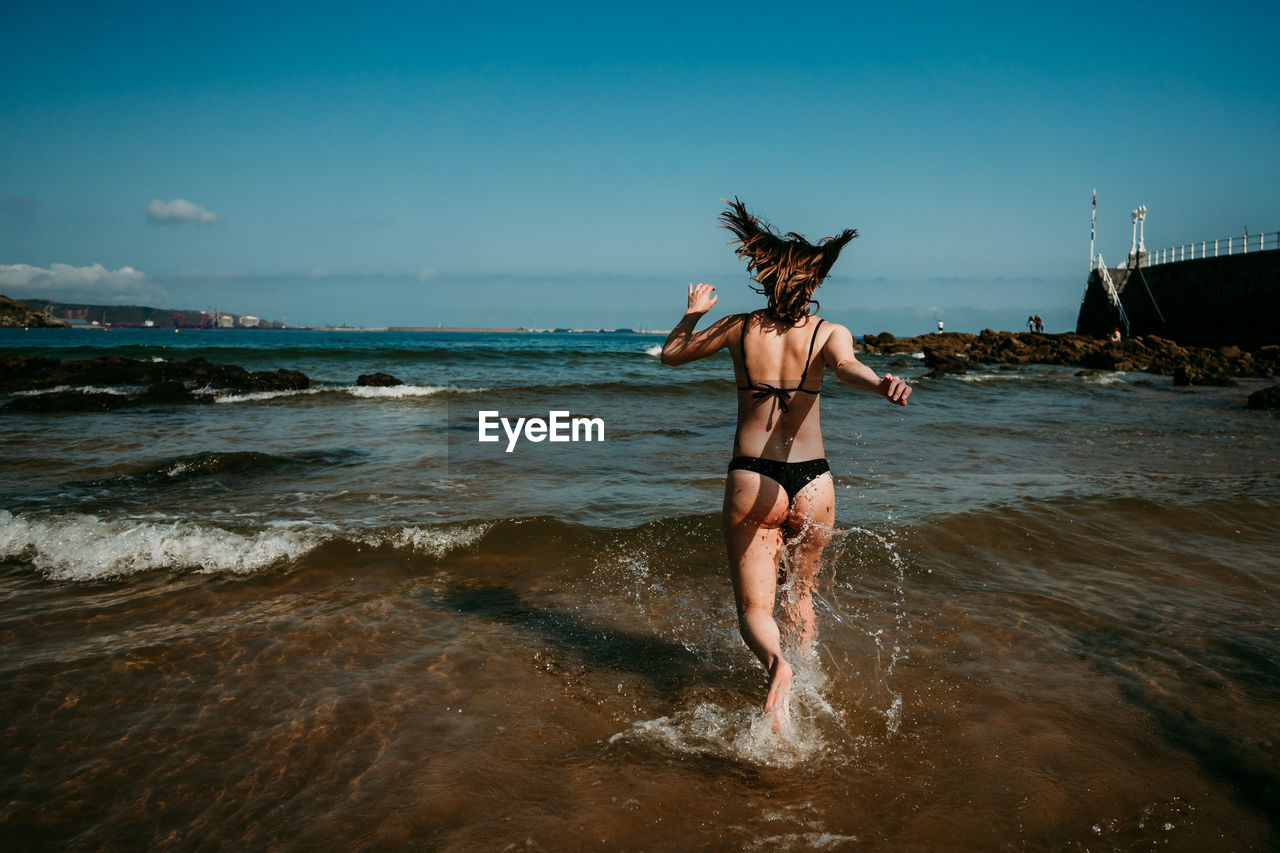 Back view of delighted female in bikini running in shallow water of sea while having fun and enjoying summer vacation