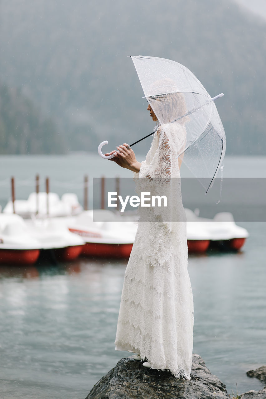 Beautiful young woman bride in a boho dress and with an umbrella stands in the rain in nature