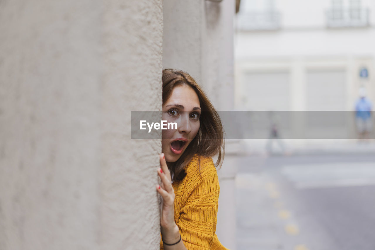 Surprised woman with brown hair peeking from wall