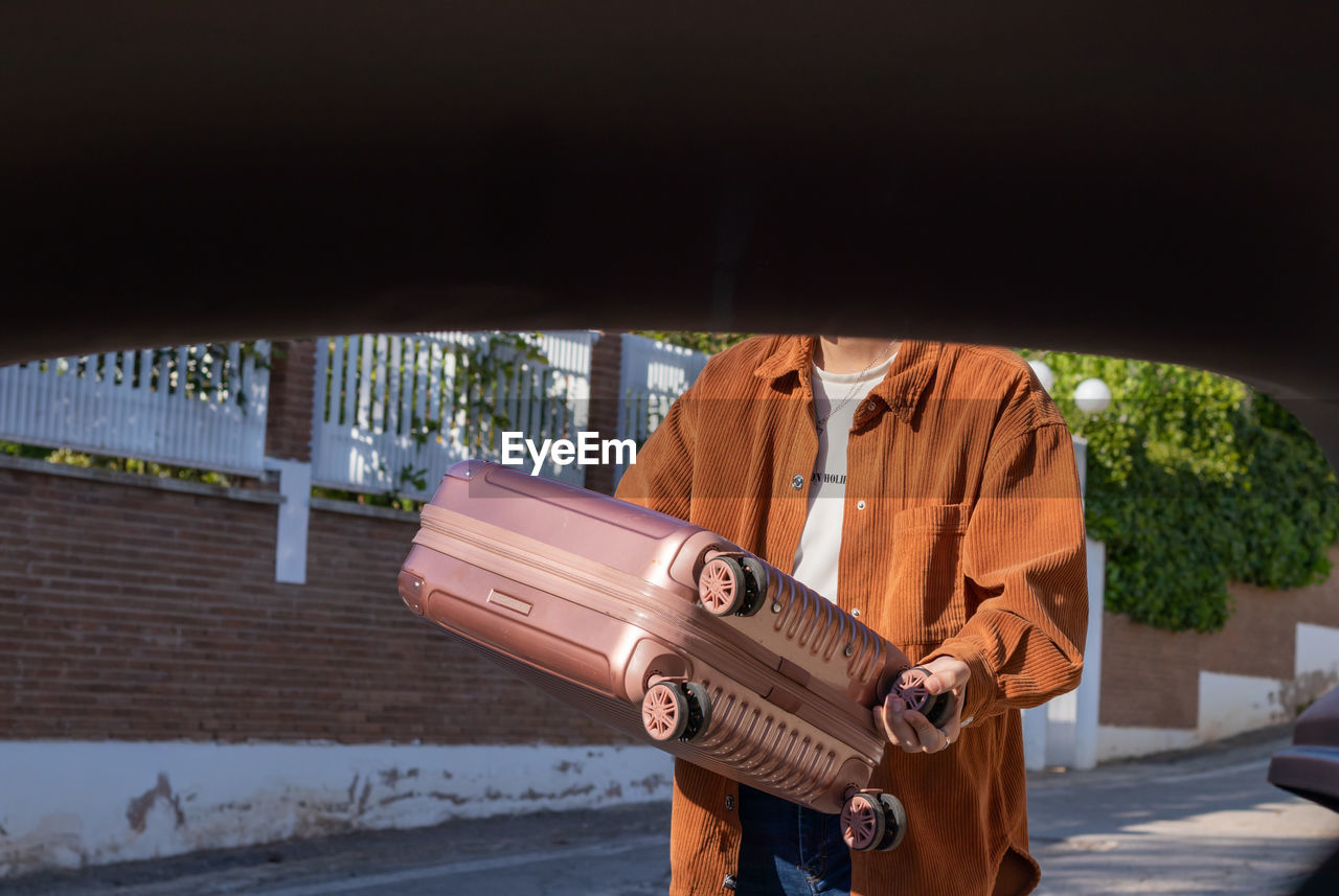 Unrecognizable young man putting carry on suitcase in the trunk of a car. view from inside the car