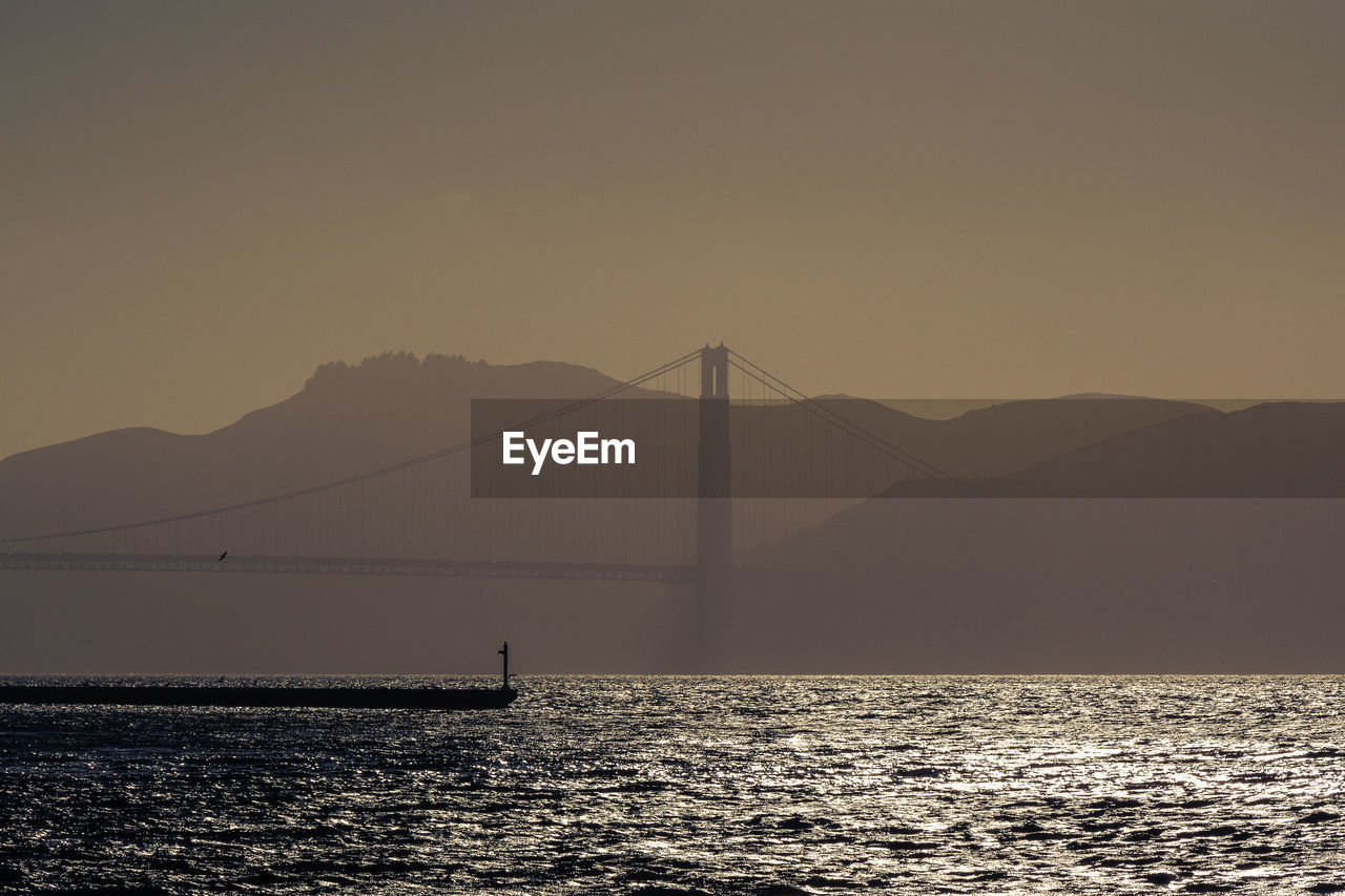 Silhouette of suspension bridge over sea against sky