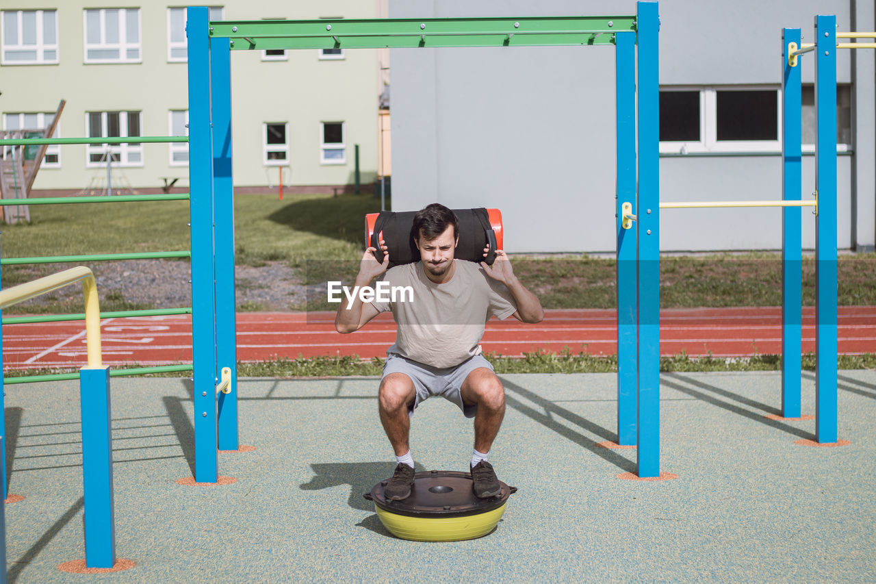 Man with a 10kg dumbbell with water on his shoulders does a squat on a balance beam