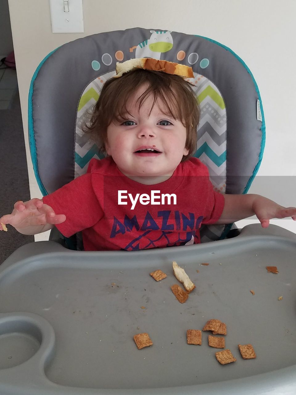 High angle view of cute boy sitting in high chair at home acting silly balancing bread on head