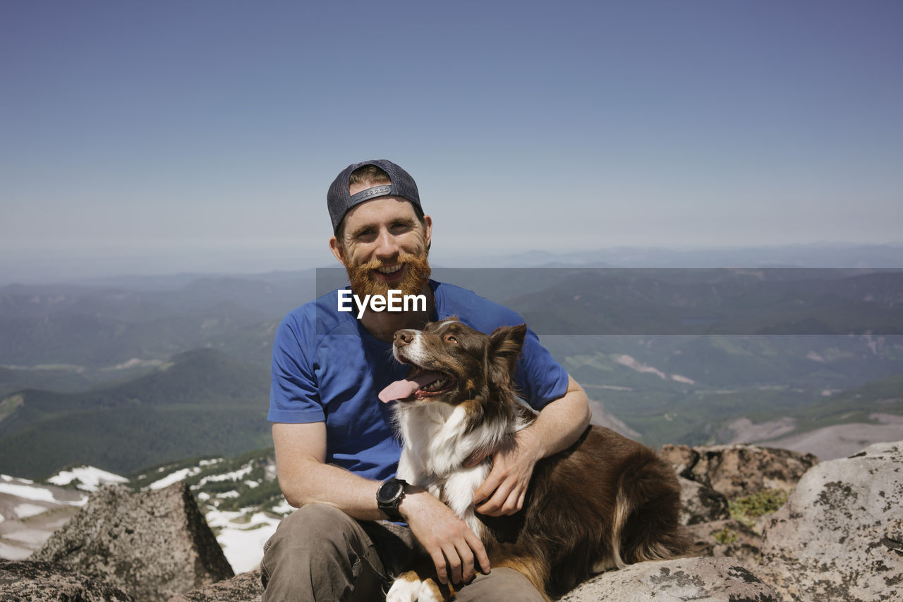 Portrait of man with dog sitting on mountain against sky
