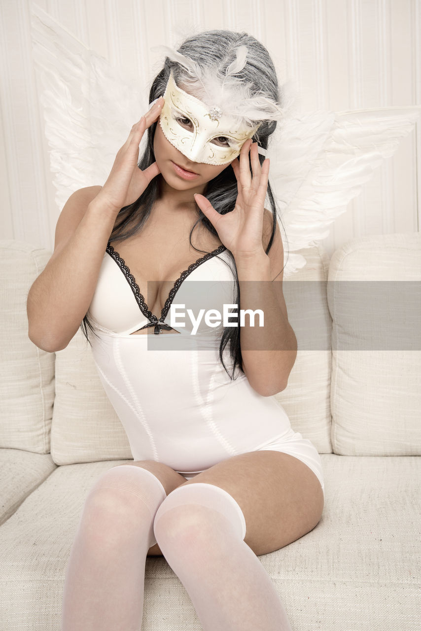 Portrait of female model in angel costume wearing eye mask on sofa