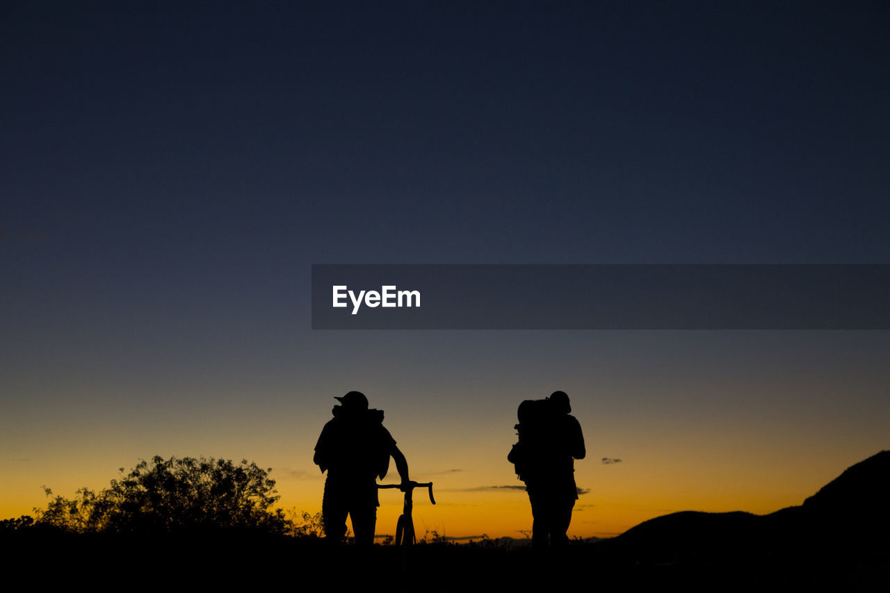 Two men with backpacks and bicycle walking into the sunset on mountain