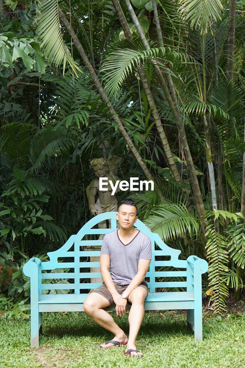 Full length portrait of man sitting on bench against trees