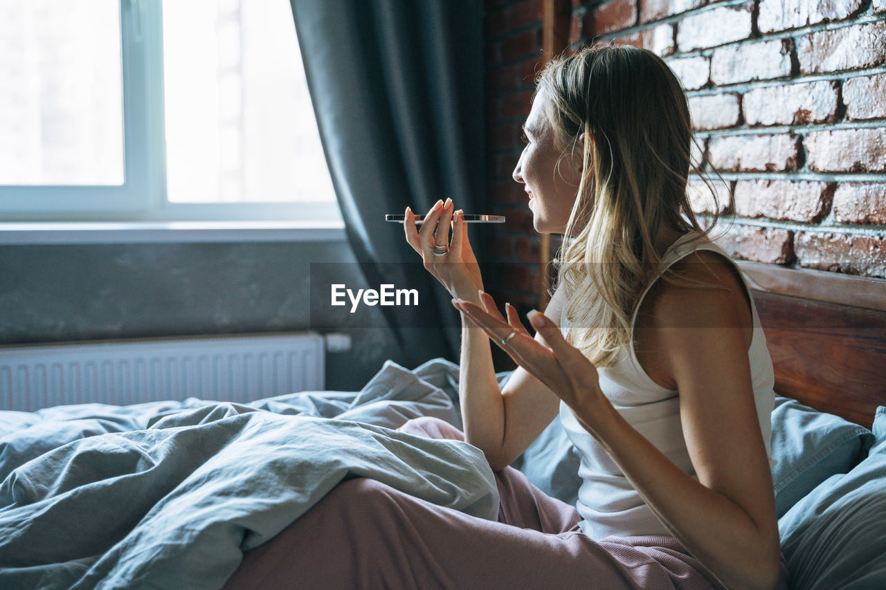 Adult woman with blonde long hair in pajamas using mobile phone on bed in loft room at home