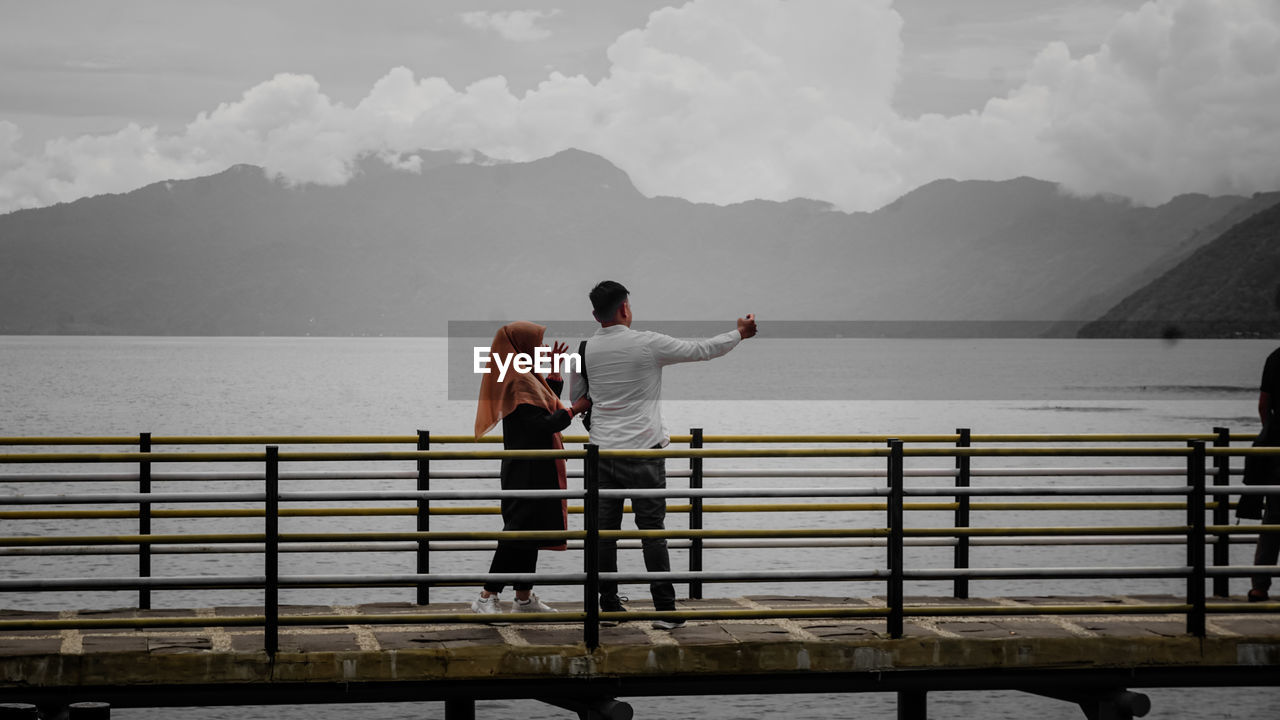 REAR VIEW OF MEN STANDING ON RAILING AGAINST MOUNTAINS