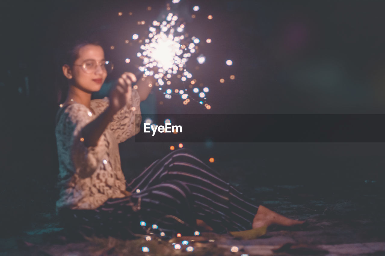 YOUNG WOMAN HOLDING SMART PHONE WHILE STANDING AT NIGHT