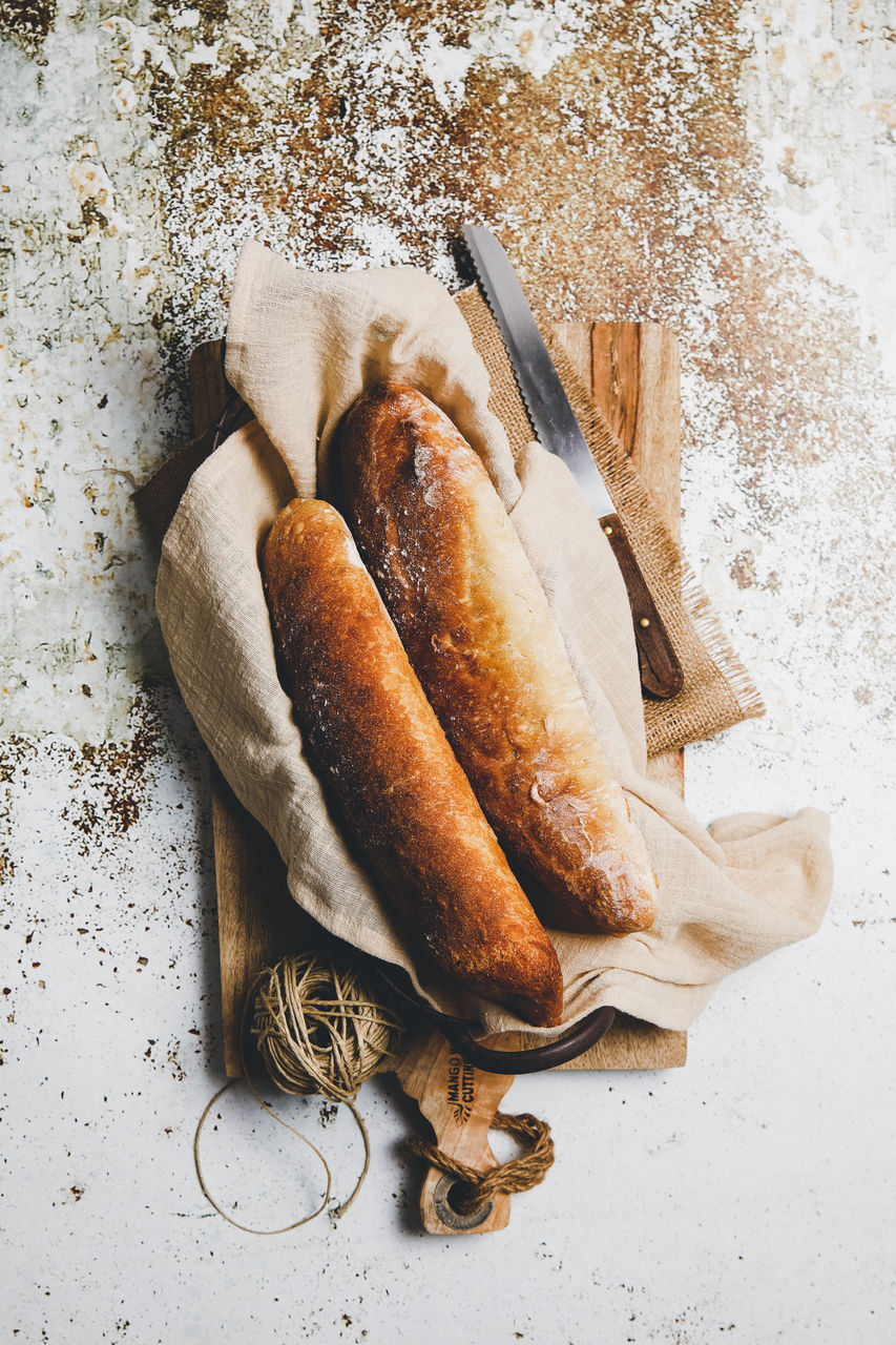 From above rustic composition with aromatic bread loaves on board with linen towel and knife on shabby surface