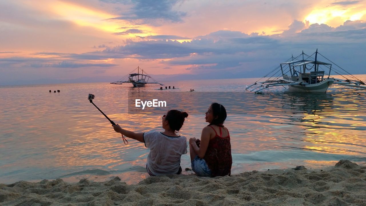 REAR VIEW OF WOMEN SITTING ON BEACH AGAINST SKY