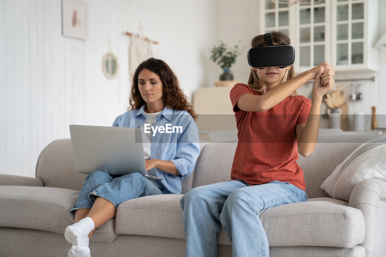 Focused teenage girl with virtual reality vr glasses holding invisible baseball bat sits on sofa