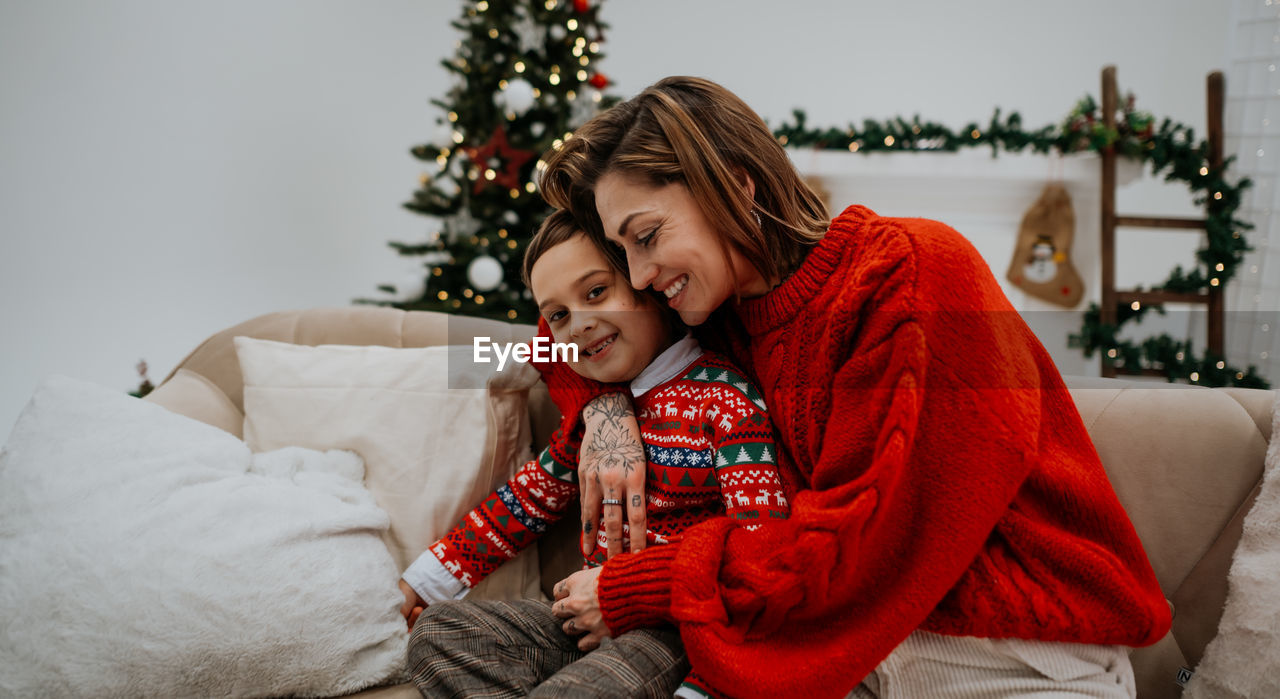 Young mother with dark hair embracing with her son near christmas tree in room decorated for holiday