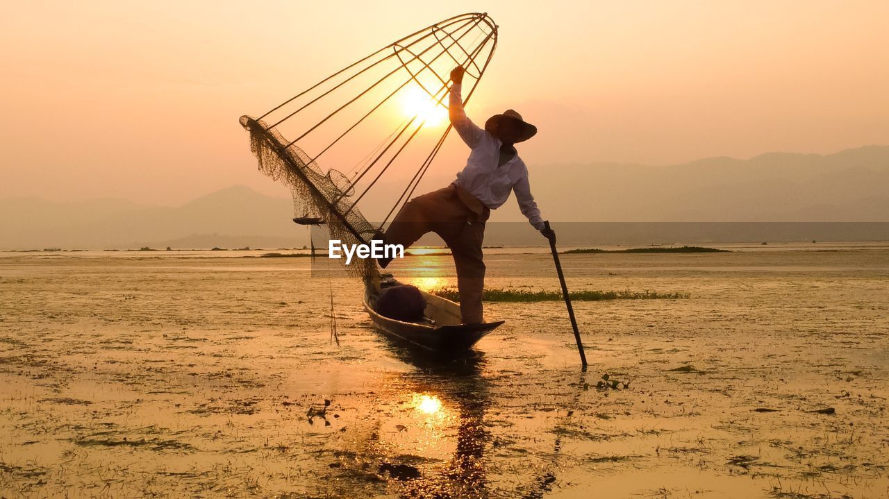 Fisherman holding fishing net and standing on boat at lake against sky during sunset