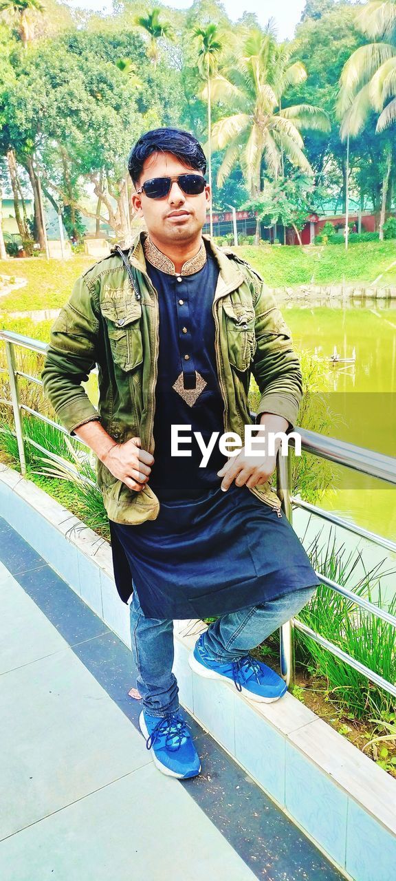 sunglasses, one person, glasses, fashion, full length, front view, adult, portrait, leisure activity, casual clothing, lifestyles, young adult, men, day, looking at camera, plant, nature, clothing, outdoors, architecture, standing, smiling, green, person, cool attitude, tree, jacket, spring, sitting