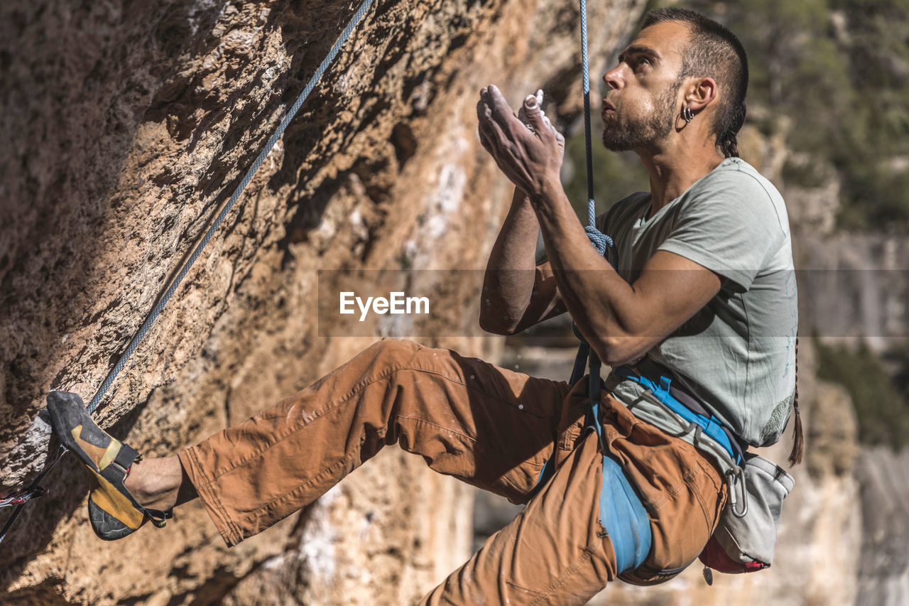 Climber talcing his hand while hanging on rope on rough cliff
