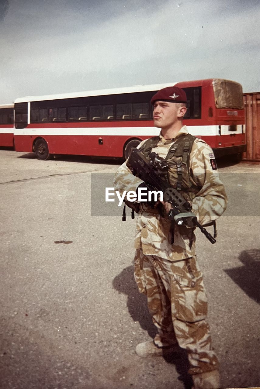 uniform, government, armed forces, military, transportation, clothing, one person, military uniform, adult, mode of transportation, soldier, standing, security, protection, army, full length, men, bus, person, occupation, war, young adult, weapon, fighting, serious, vehicle, headwear, camouflage clothing, gun, architecture