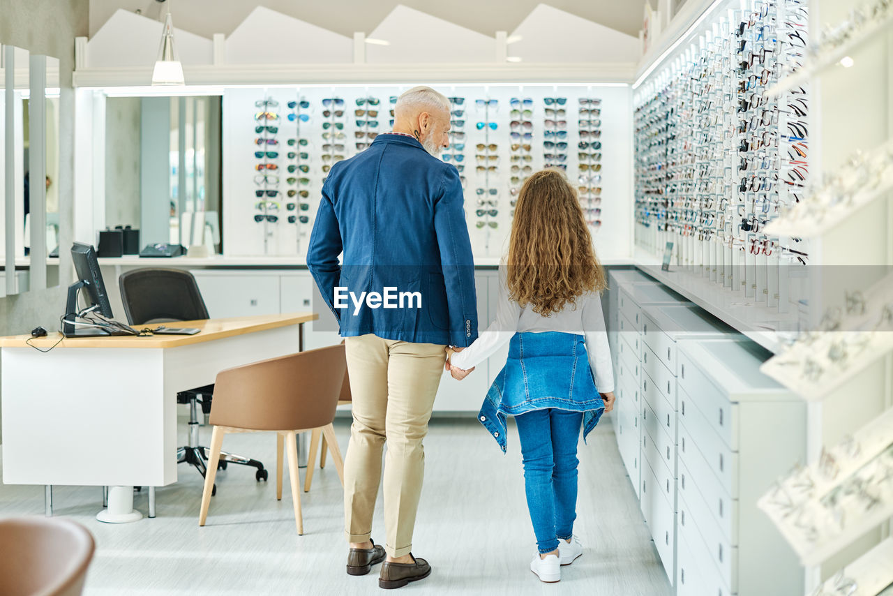 Back view of stylish senior man holding hand of teenage granddaughter while standing in optical store