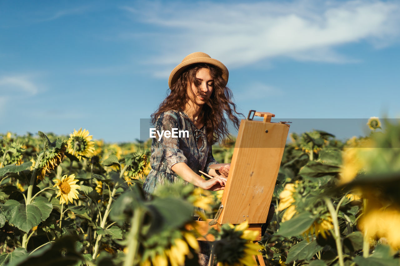 A young woman with curly hair and wearing a hat is painting in nature. 