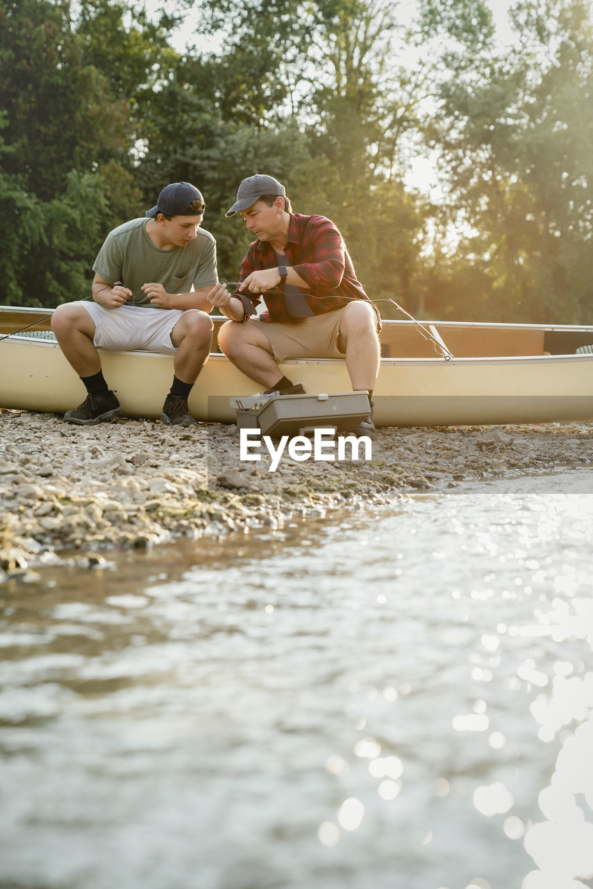 Male friends adjusting fishing rods while sitting on boat by lake