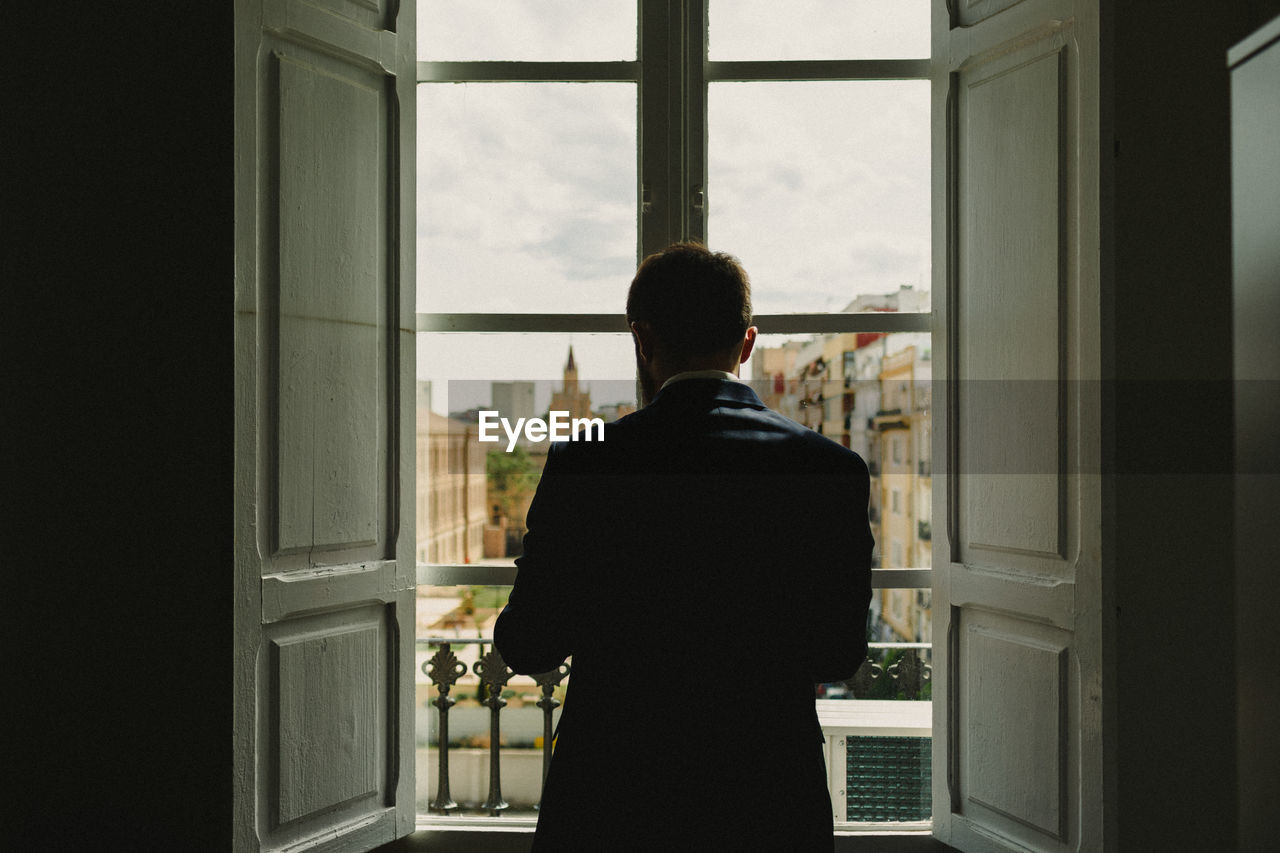 REAR VIEW OF MAN LOOKING THROUGH WINDOW AT HOME