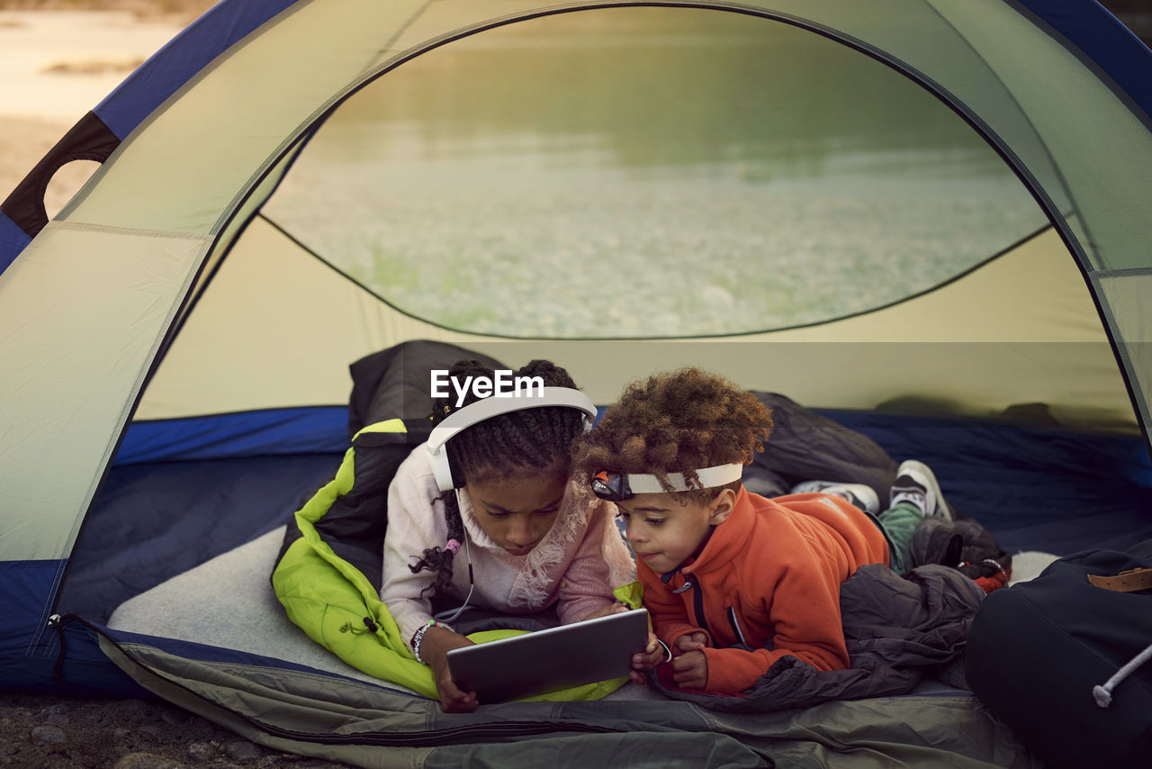 Siblings with digital tablet lying in tent