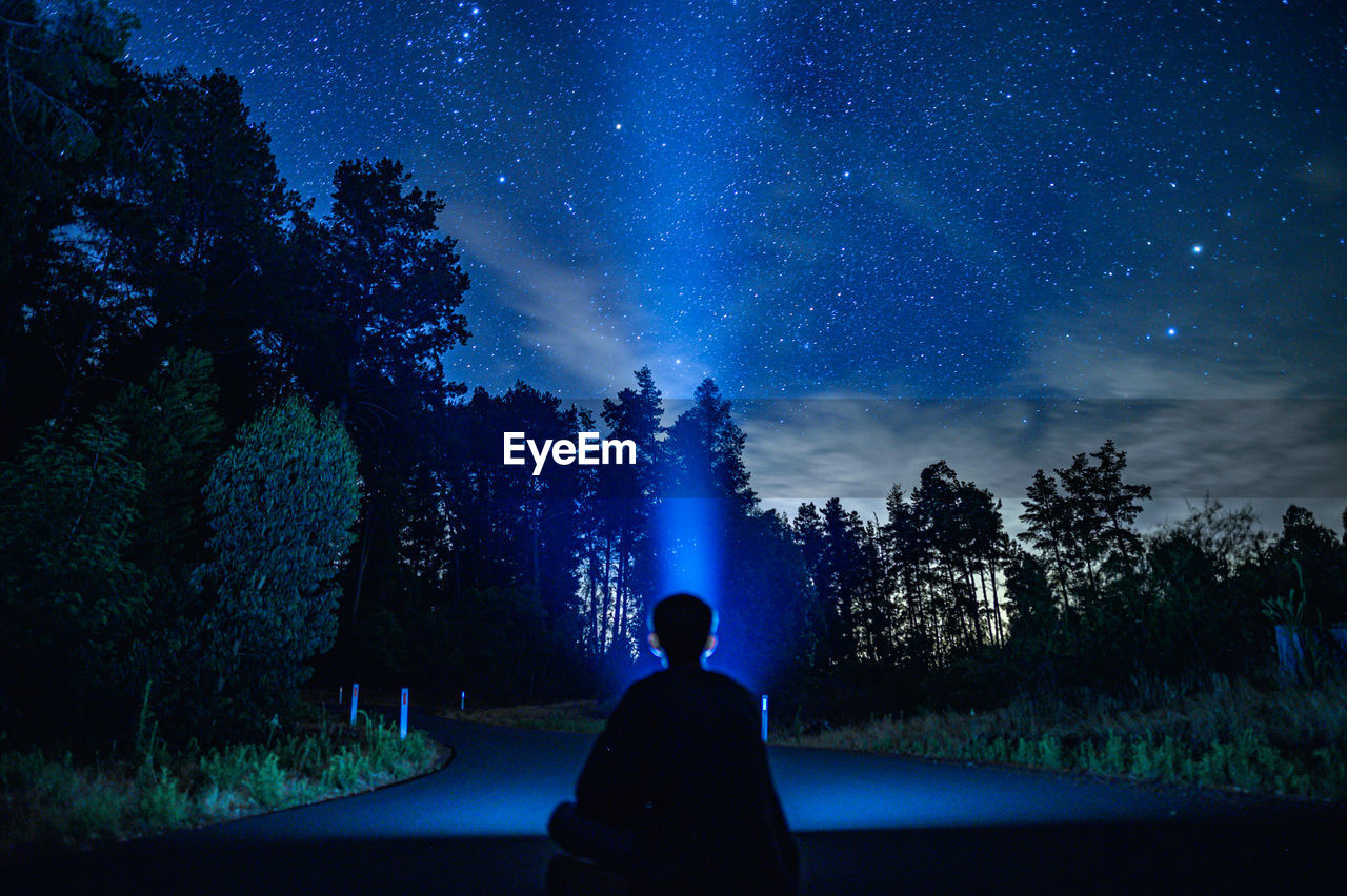 Rear view of person holding flashlight sitting against trees at night