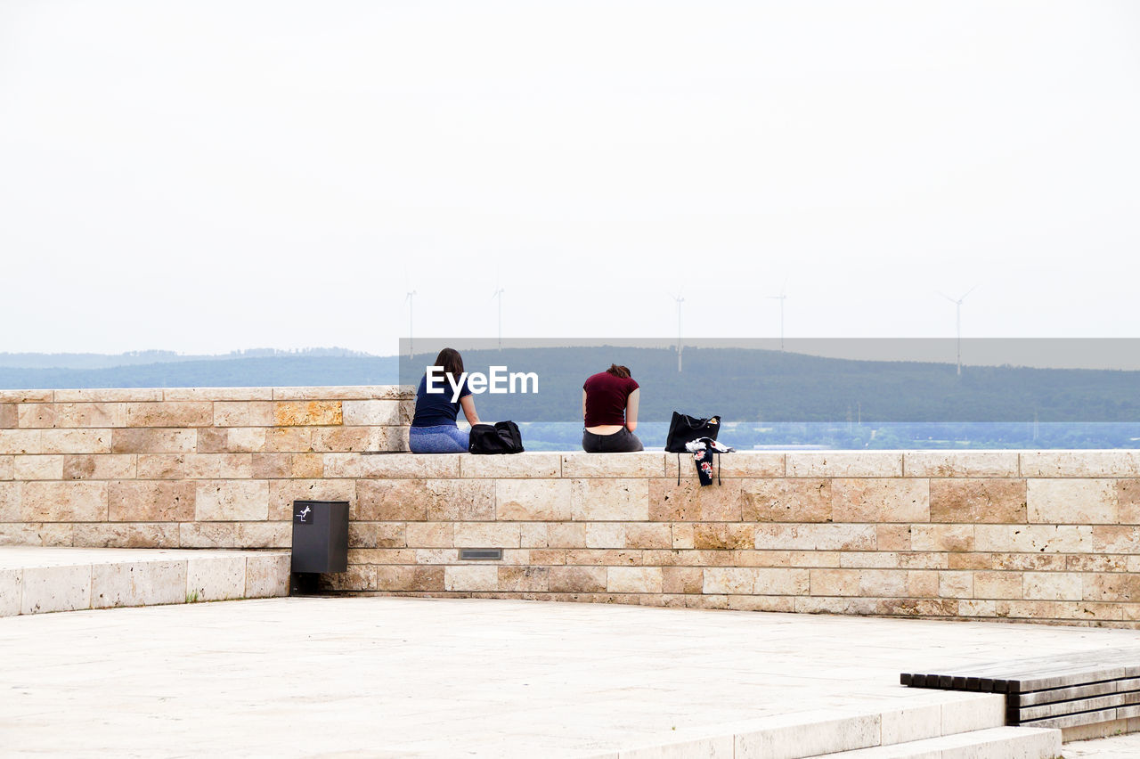 Rear view of people sitting on wall against sky