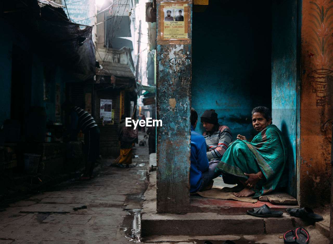Varanasi, india - february, 2018: side view of aged poor woman looking at camera while sitting with other homeless people next to old dilapidated building on narrow city street