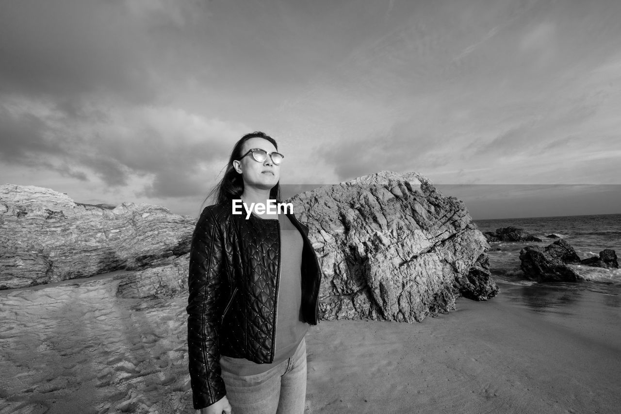 YOUNG WOMAN STANDING ON ROCK AT SEA AGAINST SKY