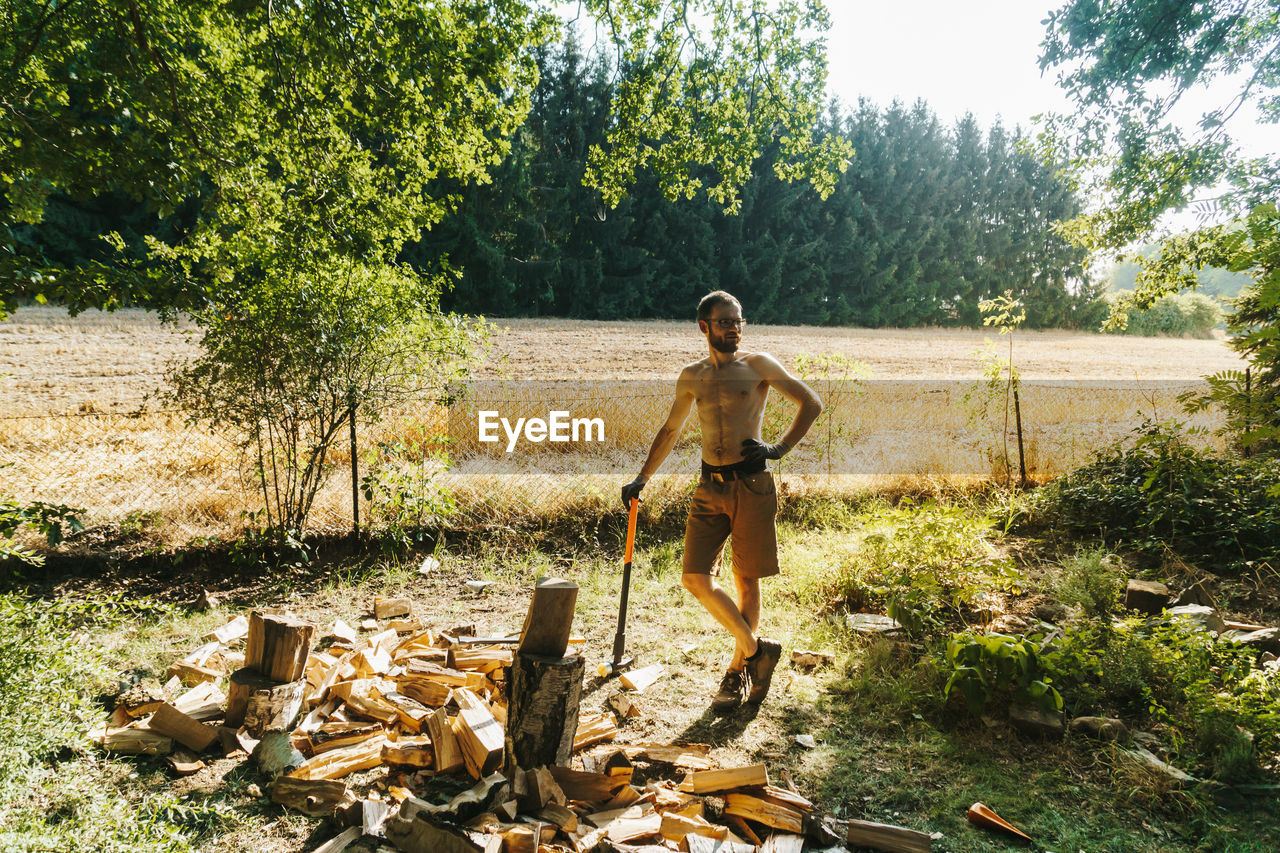 Full length of shirtless lumberjack standing by wood on field