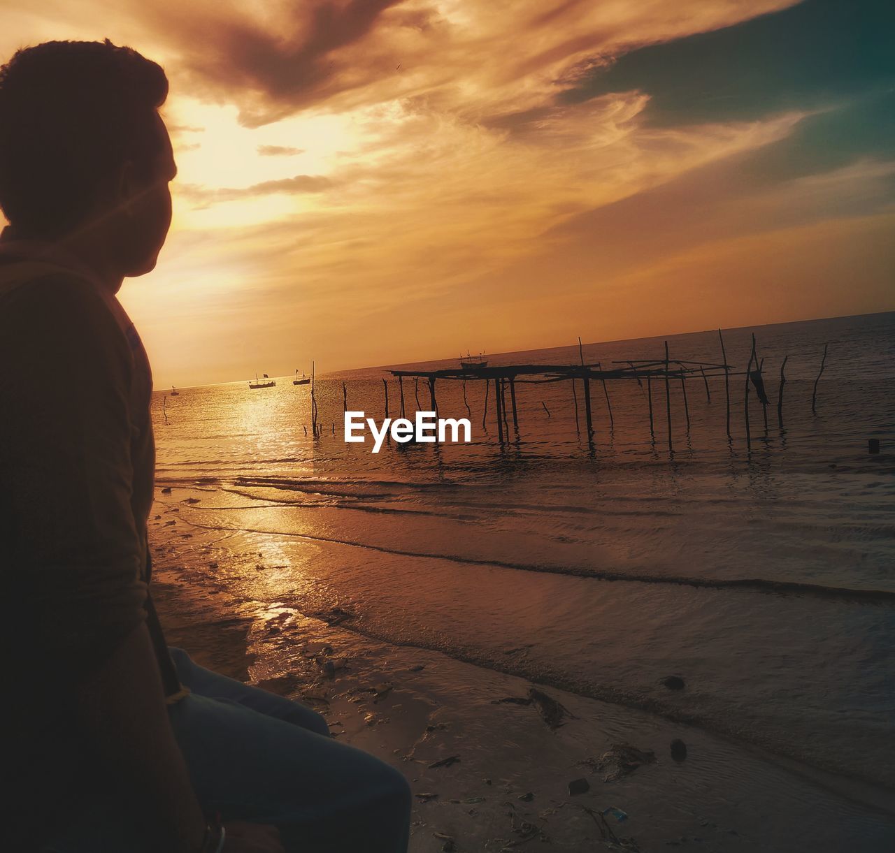 Young man sitting at beach against sky during sunset