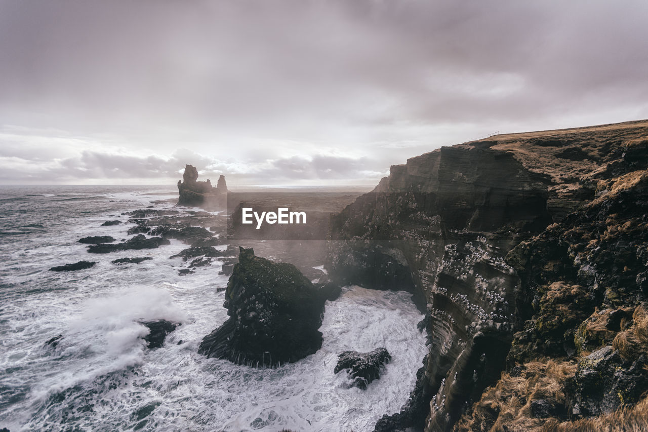 Wave splashing on cliff against cloudy sky